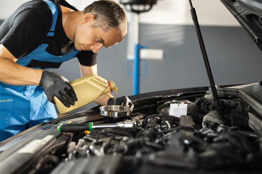 The mechanic is pouring oil into the engine. Pouring fresh oil to car engine, oil change to auto. Caucasian man in blue overalls pouring oil from plastic container