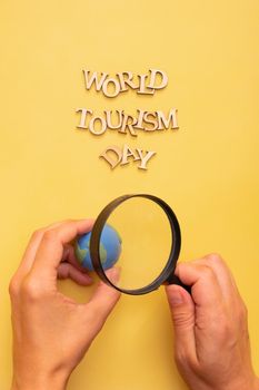 World Tourism Day text from wooden letters on a yellow color background with globe in female hands and a magnifying glass