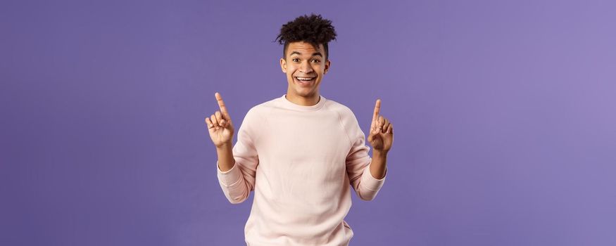 Portrait of charismatic lively young man heard about good deal, asking question intrigued, pointing fingers up, smiling upbeat at camera, look with interest, recommend product, purple background