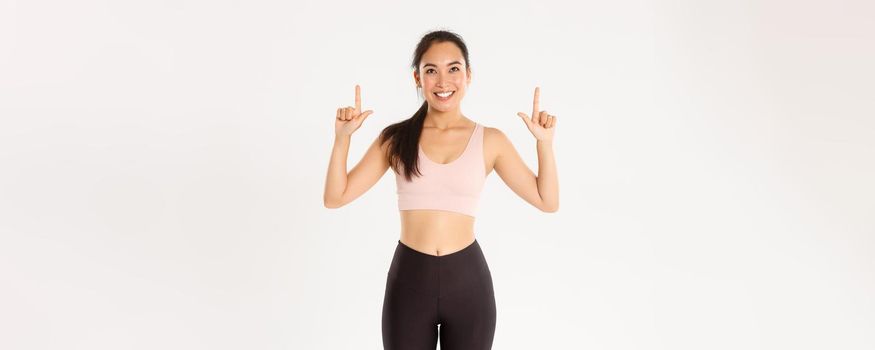 Sport, wellbeing and active lifestyle concept. Cheerful good-looking asian sportswoman in sportsbra and leggings pointing and looking up, smiling pleased, found good promo offer