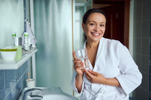 Charming woman in white bathrobe, smiling a cheerful toothy smile standing with a glass of mineral water in the bathroom