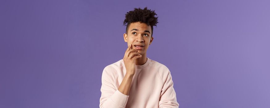 Waist-up portrait of thoughtful unsure young hispanic male student trying solve exercise, looking up thinking, pondering and calculating in mind, standing focused and indecisive purple background