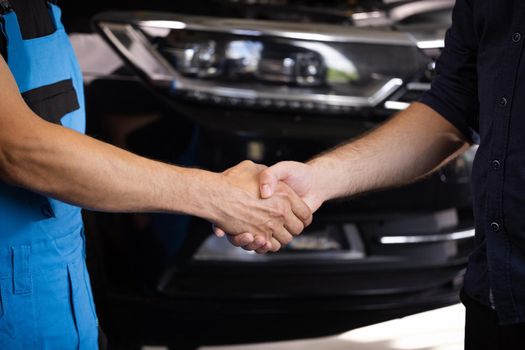 Mechanic and customer shaking hands in an auto repair shop, car service. Two unrecognizable men auto mechanic and client shaking hands at the background of vehicles. Car business, repair services