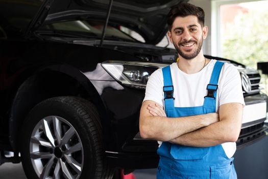 Portrait of bearded car mechanic crosses hands in a car workshop in blue uniform with equipment looking into camera. Male car mechanic at workplace in spacious repair shop