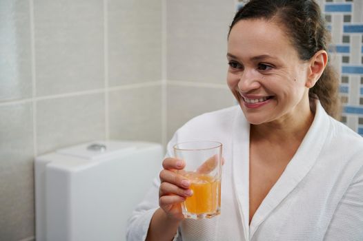 Middle-aged woman in bathrobe holds a glass of water with effervescent tablet, takes daily dose of vitamins and minerals