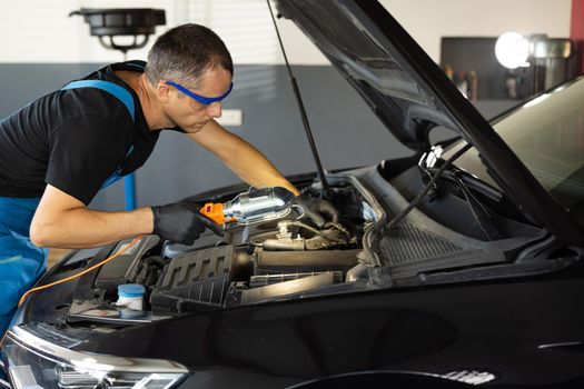 Young caucasian man in blue overalls and safety glasses inspects engine with flashlight. Male car mechanic at work in spacious repair shop. Modern workshop