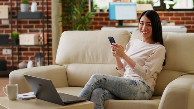 Beautiful woman having smartphone while working remotely and sitting on sofa at home