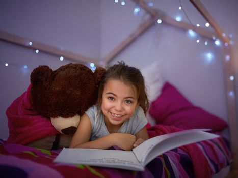 Reading lights up her imagination. Portrait of a little girl reading a book in bed with her teddybear.
