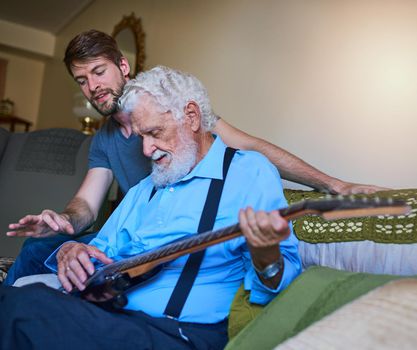 Teaching him a riff or two. a happy young man teaching his elderly grandfather to play the electric guitar on the couch at home.