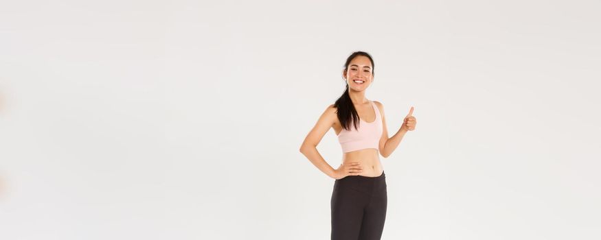 Full length of satisfied smiling, cute asian fitness girl, sportswoman in active wear showing thumbs-up and smiling pleased, proud female athelte gain daily workout goal, white background