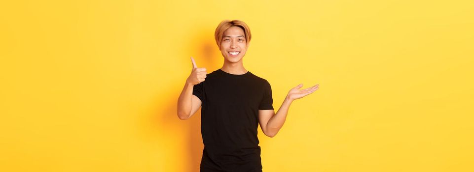 Satisfied and happy attractive korean guy smiling, showing thumbs-up in approval with rejoice, holding something on hand, standing yellow background
