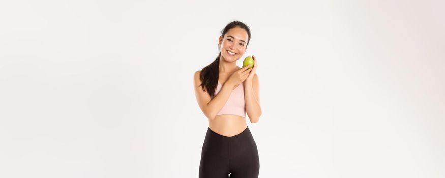 Sport, wellbeing and active lifestyle concept. Smiling cute asian fitness coach, girl athlete in sportswear showing green apple, sit on diet and workout to gain perfect body, white background