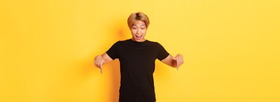 Portrait of excited and happy asian guy with blond haircut, pointing and looking down amused, standing yellow background.
