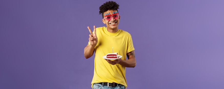 Celebration, party and holidays concept. Portrait of lovely charismatic hispaic guy in b-day glasses, celebrating his day, show peace sign photographing with birthday cake and lit candle, smiling