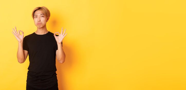 Impressed asian stylish guy with blond hair, showing okay gesture and praise something good, standing yellow background