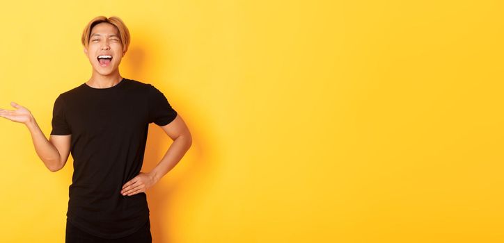 Portrait of happy blond asian guy, winking sassy and smiling, holding something on hand over yellow background