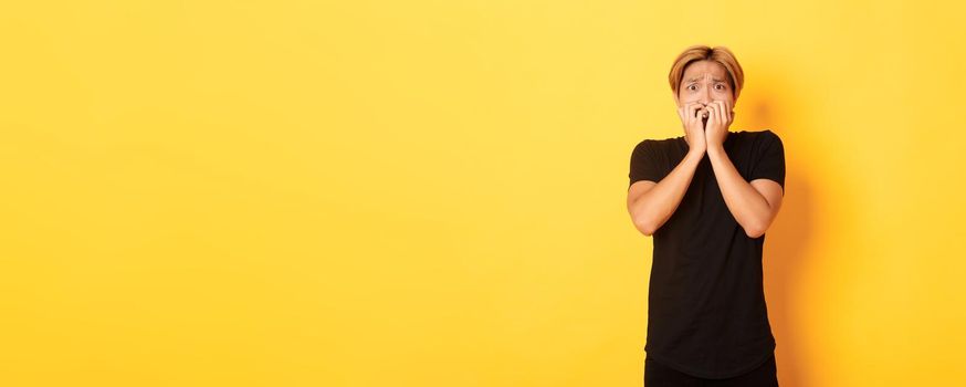 Portrait of scared insecure asian blond guy, holding hands over mouth horrified, looking frightened, yellow background