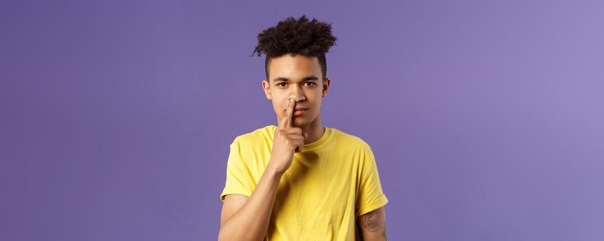 Close-up portrait of funny and unappropriate young hispanic teenage guy picking nose with finger, smiling and looking at camera, standing purple background, have bad habit