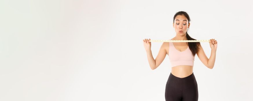 Fitness, healthy lifestyle and wellbeing concept. Impressed and happy asian female athlete showing measuring tape after measure waist and losing weight with workout program, going in gym