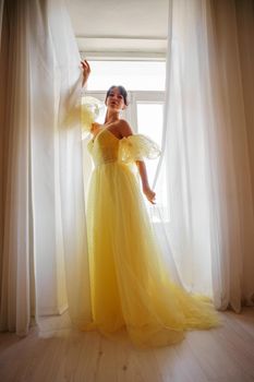 A woman's silhouette in a golden luxurious dress against the background of a window holds a curtain with her hands. Elegant lady in a yellow long silk dress with bare back, back view.