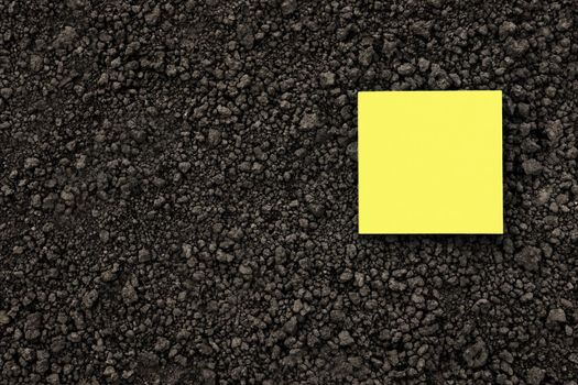 Yellow note paper square on black soil texture ground close up. Yellow sticky note blank paper flat soil organic earth texture. Mockup on ground soil background top view from above. Agriculture. Humus
