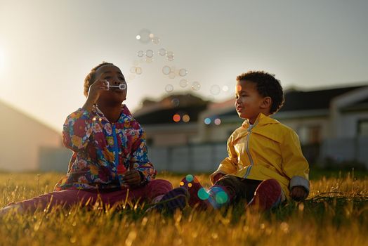 Enjoying a bit of bubble fun together. a brother and sister sitting on the ground outside blowing bubbles.