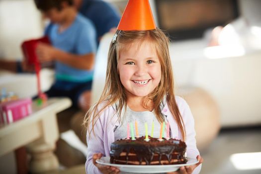 Birthday cake is the best kind of cake. Portrait of a happy little girl holding a birthday cake with her family in the background.