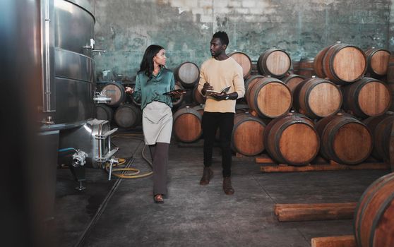 Wine cellar, warehouse and winery worker teaching a man about wine while he is writing notes. Winemaker walking in a distillery with wooden barrels and agriculture machines while consulting a partner