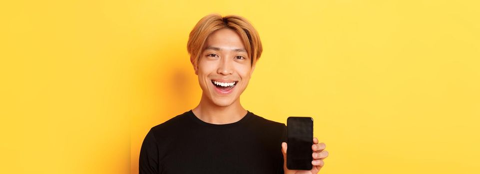 Close-up of stylish handsome korean guy showing smartphone screen and smiling pleased, recommend mobile app, standing over yellow background