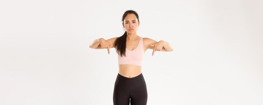 Sport, wellbeing and active lifestyle concept. Upset and disappointed asian sportswoman, woman in fitness clothing pointing fingers down with sulking face, complaining on banner, white background
