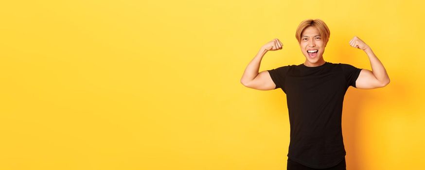 Portrait of happy and confident asian blond guy, flexing biceps, showing strong big muscles, standing yellow background