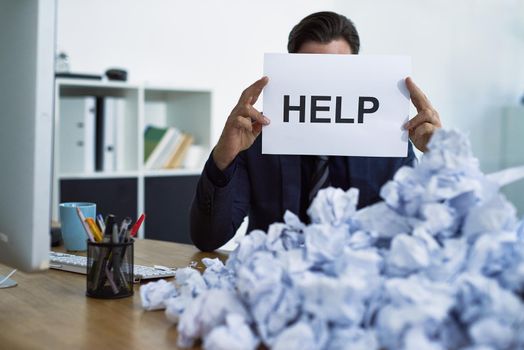 Assistance please. a stressed out businessman sitting at his desk overwhelmed by paperwork holding up a help sign.