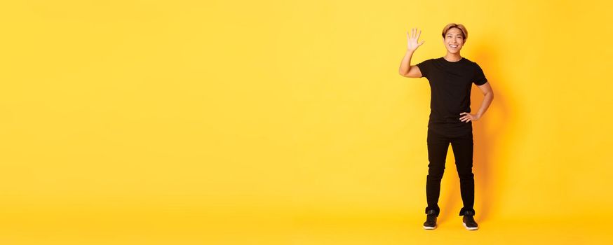 Full-length of friendly handsome asian man waving hand in hello, smiling and saying hi over yellow background