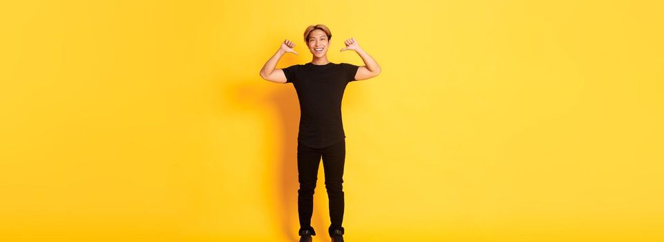 Confident attractive blond asian guy, smiling sassy and pointing at himself, standing over yellow background