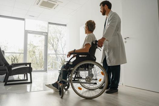 Professional male doctor carrying female patient on wheelchair in medicine clinic hall