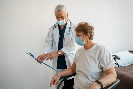 Doctor in mask explaining to a sick patient in a wheelchair the details of treatment