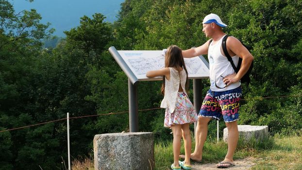 beautiful park in alpine mountains. man and a girl, kid, tourists, study map of park, against backdrop of picturesque landscape of the Alpine mountains and a small town. summer hot day