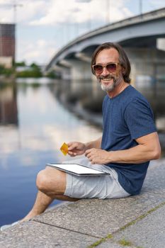 Confident handsome freelancer man hold credit, debit card sitting outdoors next to river working using digital tablet pc, shopping online, paying online banking. E-banking concept. Financial concept