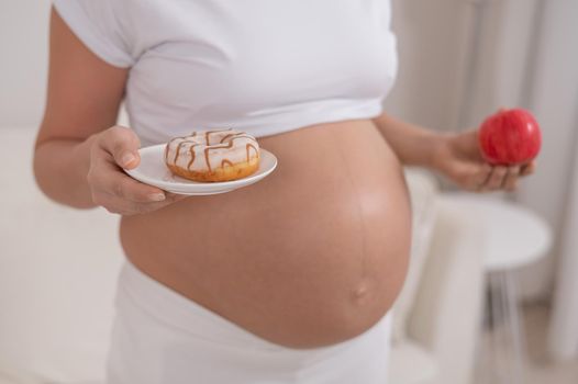 A pregnant woman is holding a red apple and donut. Choice of food.