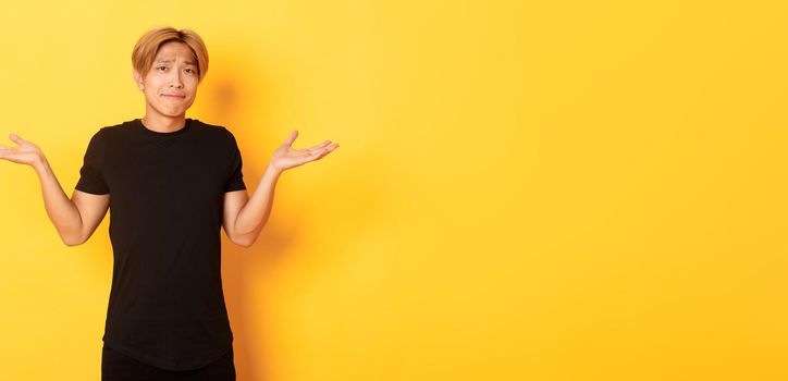 Portrait of confused and indecisive attractive korean guy, shrugging and pouting upset, standing yellow background