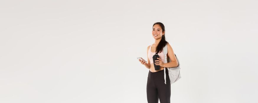 Full length of smiling healthy, slim asian girl going fitness training, female athelte carry backpack with workout equipment and water bottle, using mobile phone sports application, white background