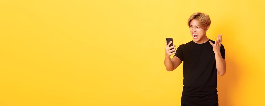 Portrait of angry and pissed-off asian man looking mad at smartphone screen, having argument during video call, standing yellow background