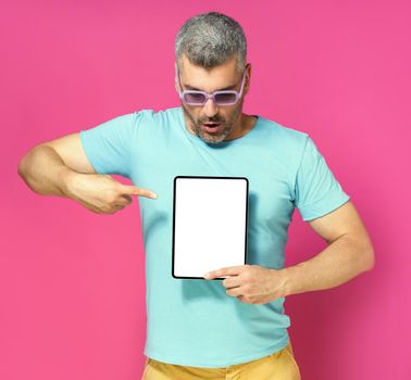 Pointing at digital tablet handsome man looking at white screen lowered his head wearing casual blue shirt and sunglasses isolated on pink background. Mobile app advertisement