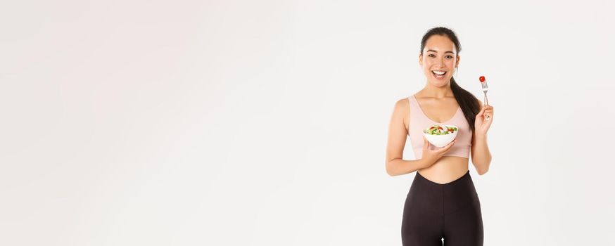 Sport, wellbeing and active lifestyle concept. Smiling slim and healthy asian girl doing fitness exercises, losing weight with gym workout and eating fresh salads, standing white background