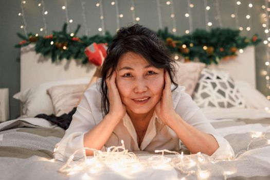 Senior woman relax on bed with xmas lights, charming elderly smiling lady in bedroom at christmas, atmospheric mood