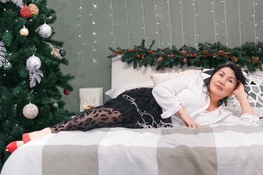 Senior full length woman rest on bed with xmas decoration and lights, elderly smiling lady dream and relax at christmas