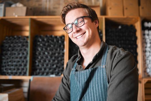 Happy, smile and proud wine, warehouse or manufacturing worker wearing glasses in a cellar with a bottle collection on display. Winery manager or employee in the alcohol industry or distillery