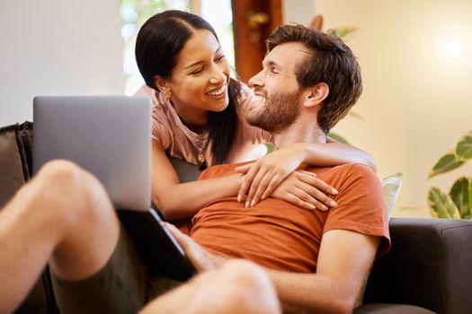 Happy couple bonding and streaming on laptop while relaxing on couch at home, talking and laughing. Interracial husband and wife enjoying their relationship, embracing and watching a movie together