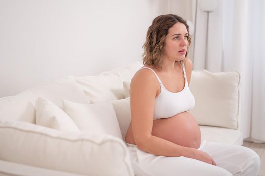Tired pregnant woman sits on a white sofa.