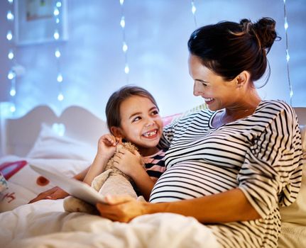 Whos picking the story tonight. an attractive young pregnant woman reading her daughter a bedtime story on a tablet.